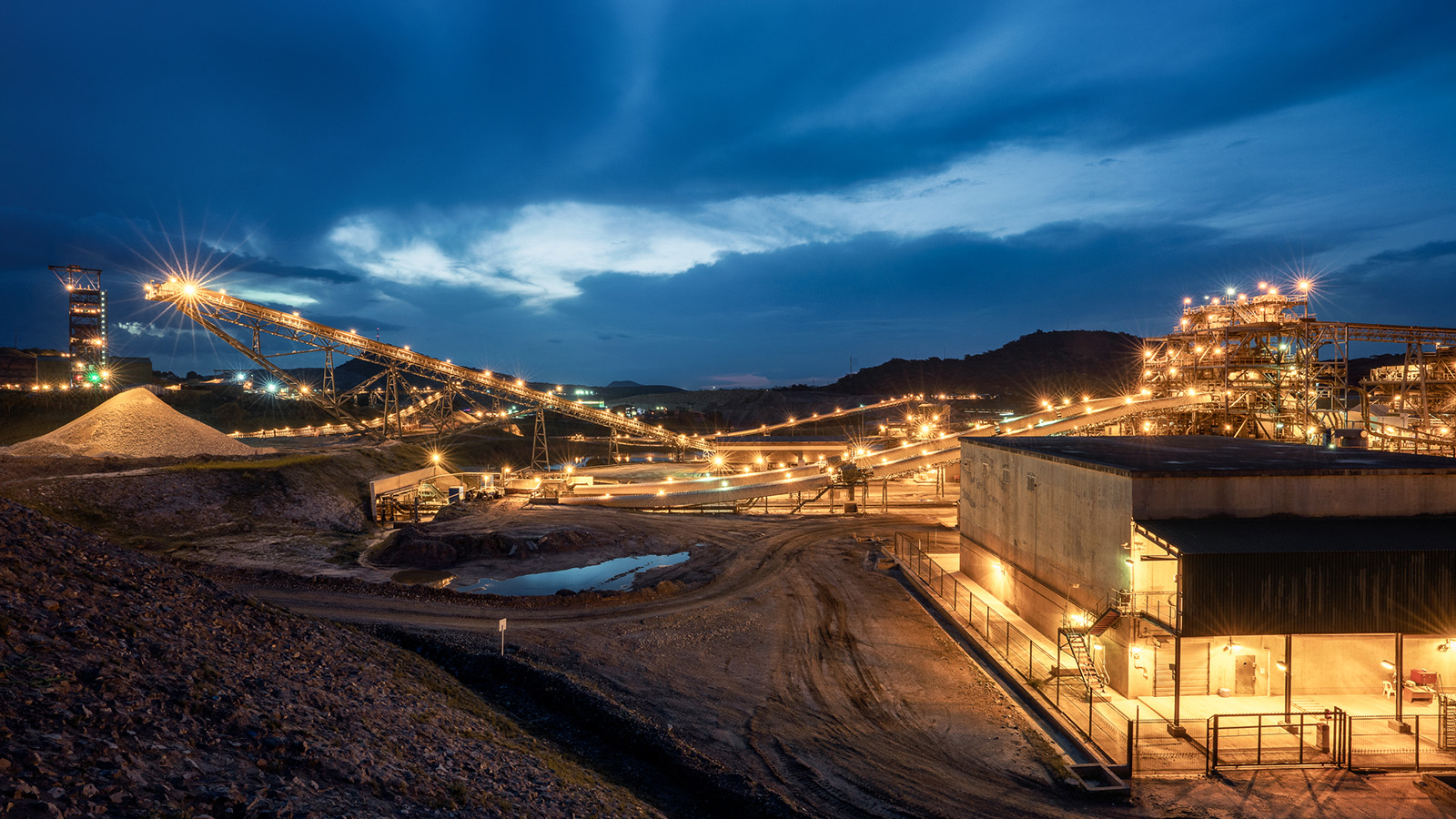 A large-scale mining operation photographed at dusk, showing illuminated industrial infrastructure including conveyor belts, processing facilities, and excavation equipment. The scene features multiple levels of conveyor systems transporting materials across the mining site, with bright industrial lighting creating star-burst effects against the dramatic blue twilight sky. Piles of extracted material are visible in the foreground, along with concrete processing buildings and mining equipment. The operation is set in a hilly, rocky landscape with mountains silhouetted in the background.