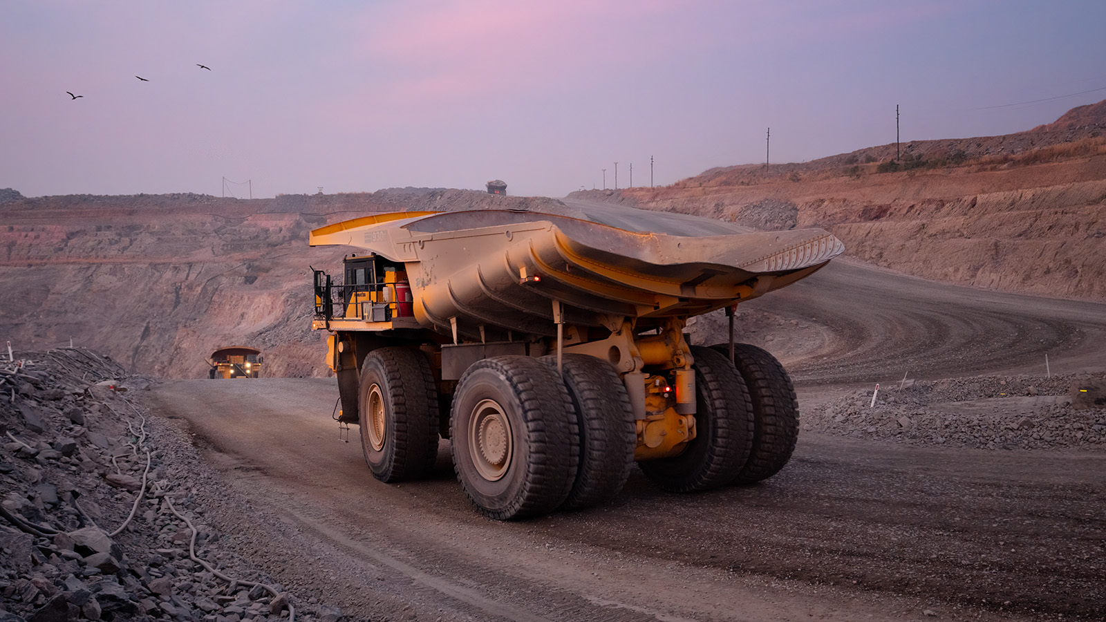  mining haul truck drives along a dirt road in an open-pit copper mine in Zambia during golden hour. The enormous dump truck, designed for heavy-duty mining operations, is illuminated by warm lighting against a backdrop of terraced mining benches carved into reddish-brown earth. A second haul truck is visible in the distance. The desert-like landscape shows the characteristic stepped excavation pattern of open-pit mining, with utility poles visible on the horizon and a few birds flying in the dusky purple sky.