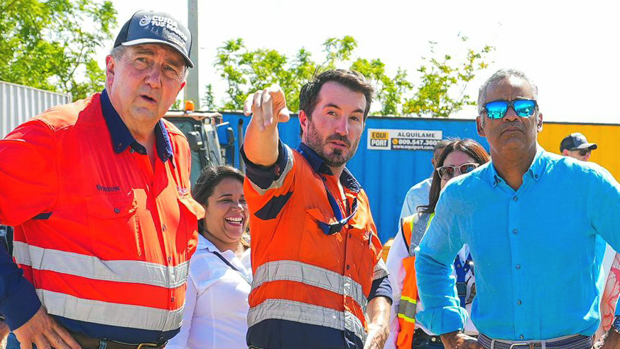 A group of officials and workers gather at a construction or infrastructure site. Several people wear high-visibility safety vests in orange and red with reflective stripes, while others are dressed in business attire including blue shirts. The scene appears to be at an official visit or project inspection, with industrial equipment and storage containers visible in the background among green vegetation.