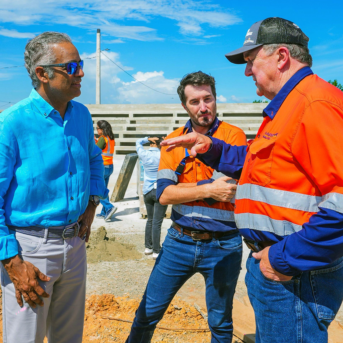 Three men engage in conversation at what appears to be a construction site, with one man in a blue business shirt and sunglasses speaking with two workers wearing orange high-visibility safety uniforms with reflective stripes and hard hats. The setting shows a concrete structure under construction with a partly cloudy sky and rural landscape in the background.