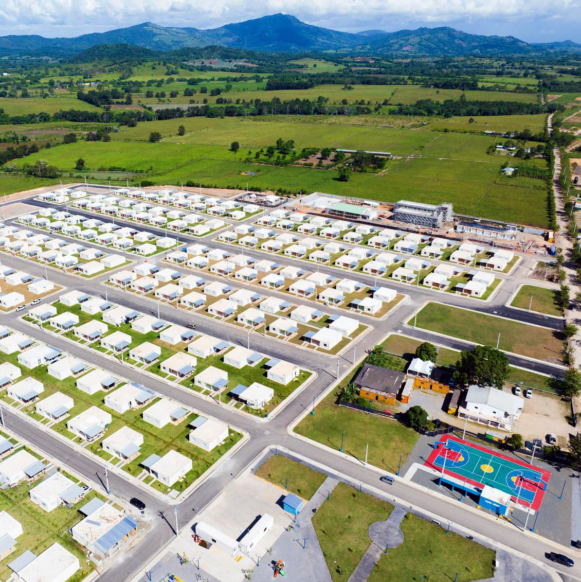 An aerial view of the New Horizons residential community showing a well-planned neighborhood layout with dozens of small, single-story white houses arranged in a grid pattern along paved streets. The development includes community amenities such as a colorful basketball court and playground equipment. The community is surrounded by lush green agricultural fields and rolling hills under a partly cloudy sky, demonstrating the rural Dominican Republic setting near the Pueblo Viejo gold mine.