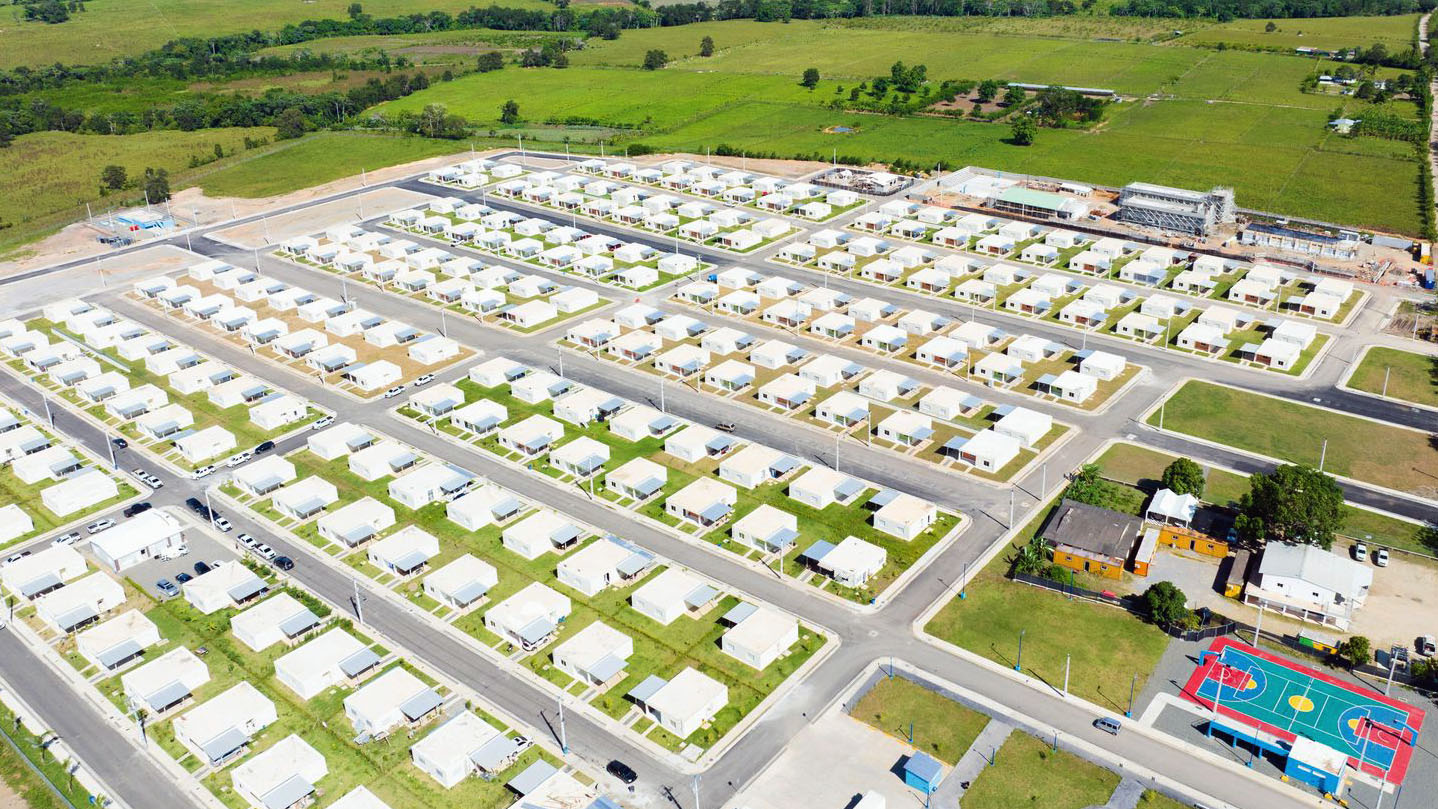 An aerial view of the New Horizons residential community showing a well-planned neighborhood layout with dozens of small, single-story white houses arranged in a grid pattern along paved streets. The development includes community amenities such as a colorful basketball court and playground equipment. The community is surrounded by lush green agricultural fields and rolling hills under a partly cloudy sky, demonstrating the rural Dominican Republic setting near the Pueblo Viejo gold mine.