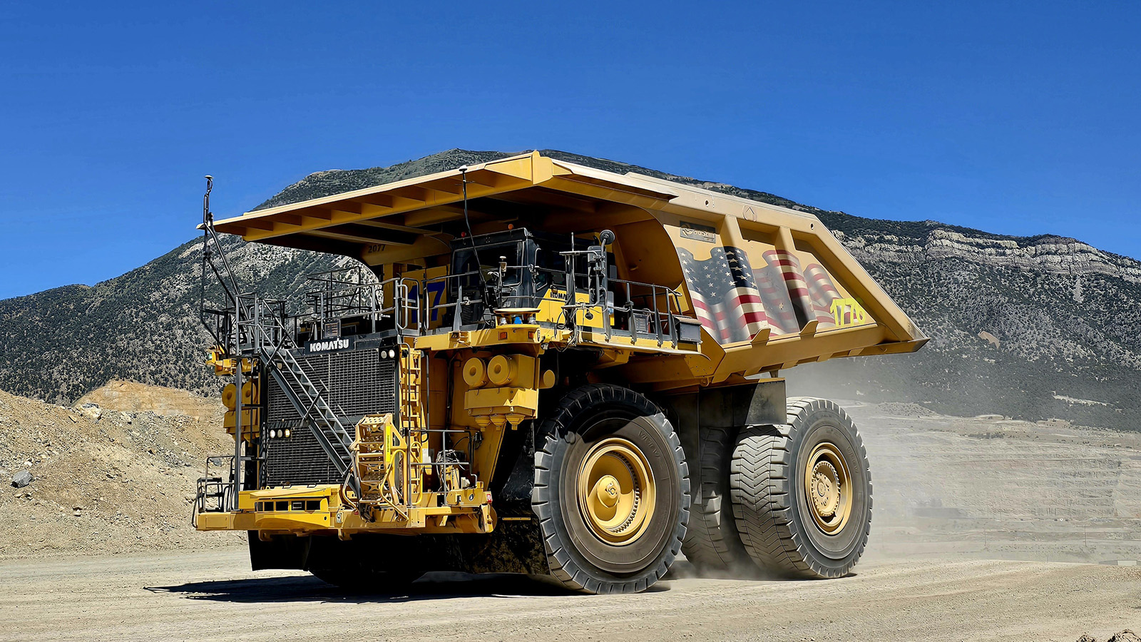 A massive yellow Komatsu haul truck with an enormous dump bed sits on a dirt road at an open-pit mining site in Nevada. The truck features huge black tires, external staircases for cab access, and appears to have an American flag design on its dump bed. Behind the vehicle, barren desert mountains rise against a clear blue sky, with exposed rock faces and mining tailings visible on the hillsides, characteristic of Nevada's gold mining operations.