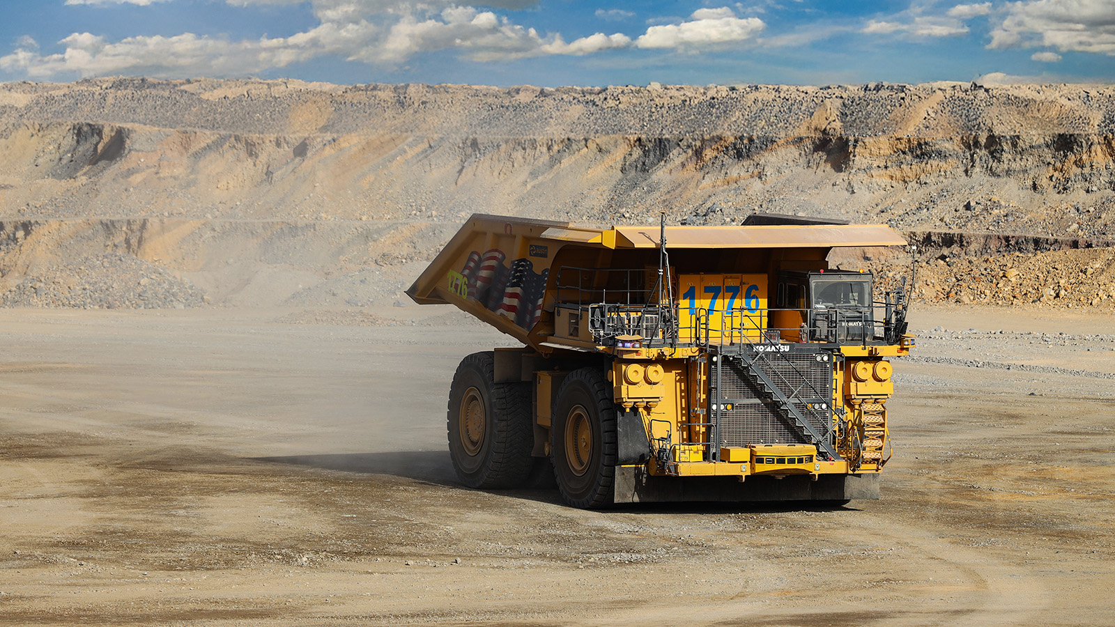 A massive yellow Komatsu haul truck numbered 1776 drives across a dirt road at an open-pit gold mine in Nevada, kicking up dust behind its enormous tires. The truck features a large dump bed with an American flag design and external metal staircases leading to the operator's cab. In the background, the dramatic terraced walls of the open-pit mine stretch across the landscape under a partly cloudy blue sky, showing the characteristic benched mining levels carved into the desert terrain.