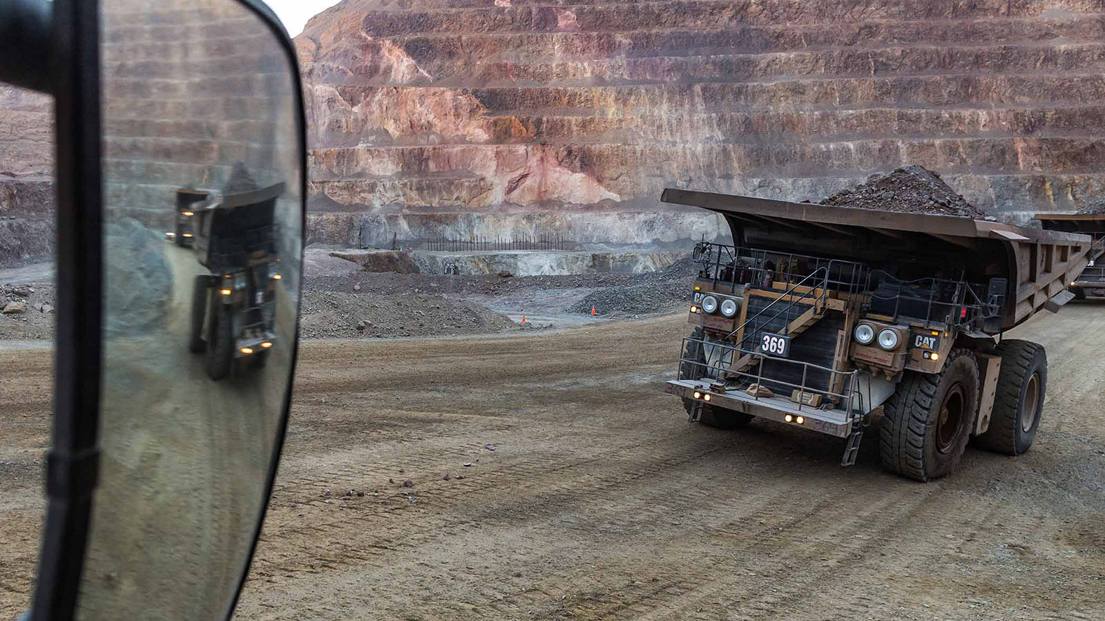 View from inside a haul truck cab showing a loaded CAT mining truck (unit 369) driving through an open-pit mine, with terraced rock walls in the background and a second truck reflected in the side mirror.