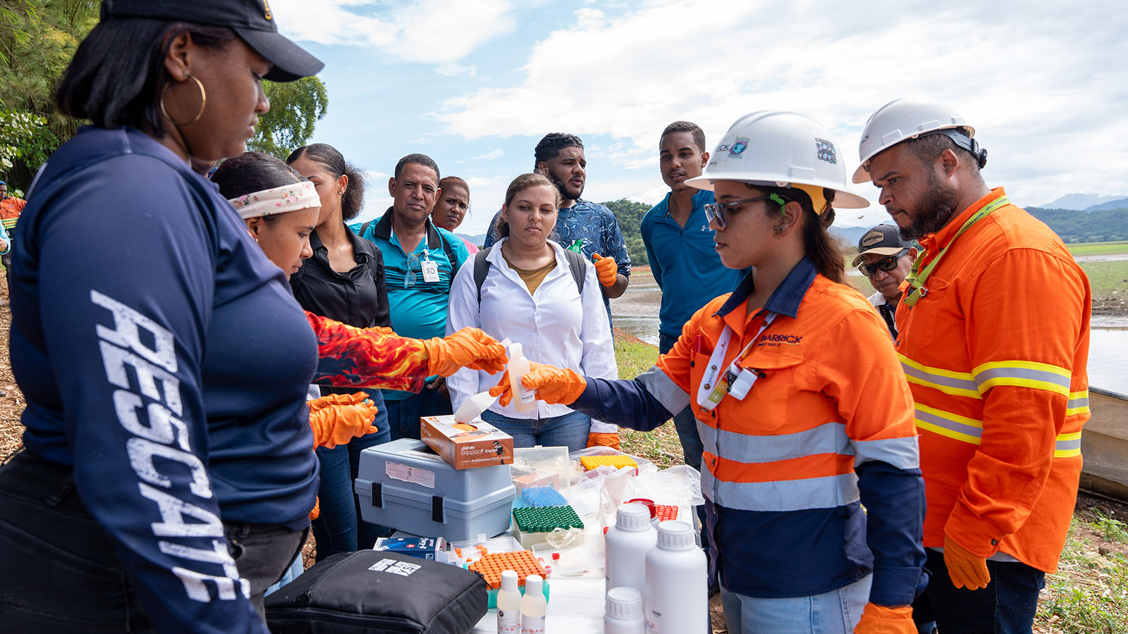 Barrick team members demonstrate water-sampling procedures to community participants during a participatory environmental monitoring activity near a waterbody. The group gathers around a field testing station equipped with sample containers, testing supplies, and protective equipment as environmental samples are prepared and collected.