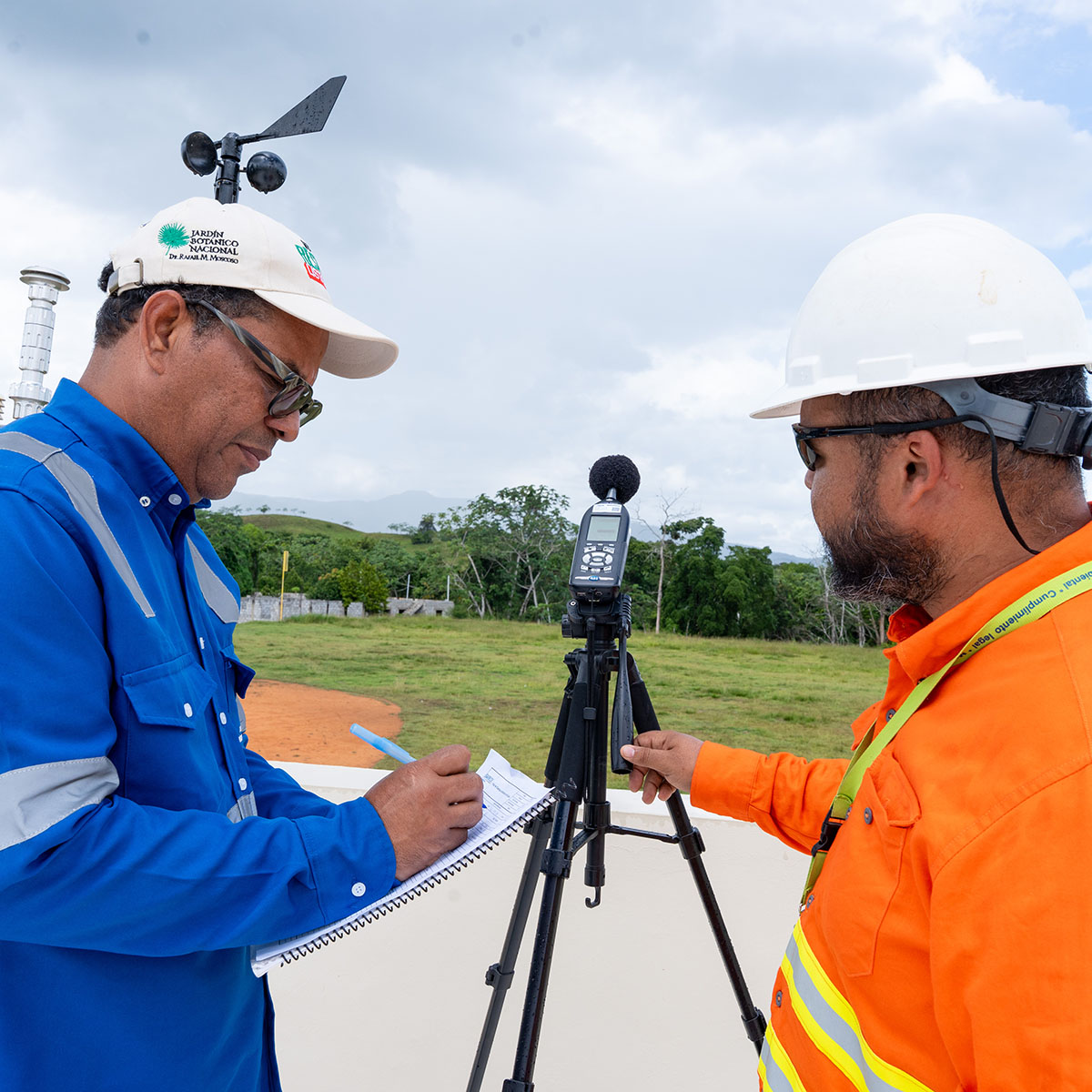 A participant and a Pueblo Viejo environmental specialist review measurements from a sound-monitoring instrument mounted on a tripod during a participatory environmental monitoring activity. Data is recorded on-site as part of community-led verification of environmental conditions around the operation.