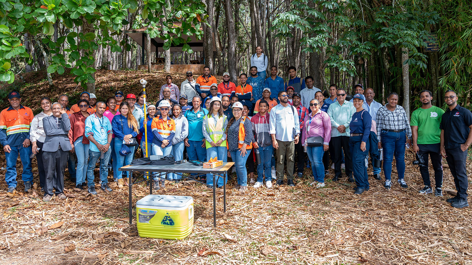 Community representatives, local stakeholders, and Pueblo Viejo team members gather for a group photo during the mine’s participatory environmental monitoring program. The participants are assembled in a wooded outdoor area alongside environmental monitoring equipment and field sampling supplies used during the monitoring activities.