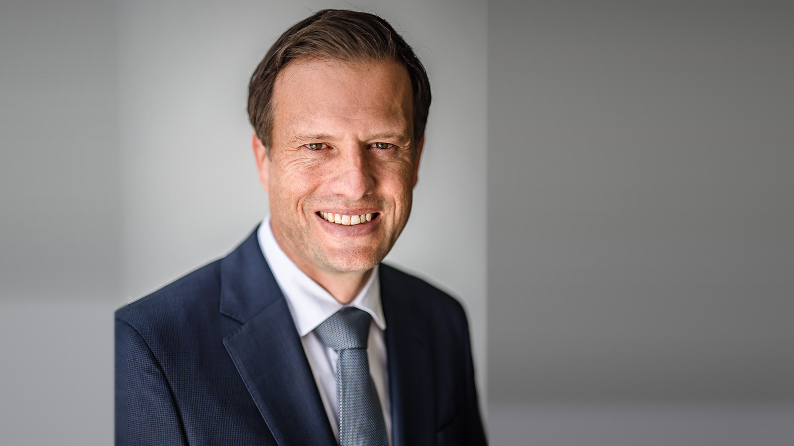 Professional headshot of Sebastiaan Bock smiling with short brown hair, wearing a dark navy suit, white dress shirt, and light blue textured tie, photographed against a soft gray background.