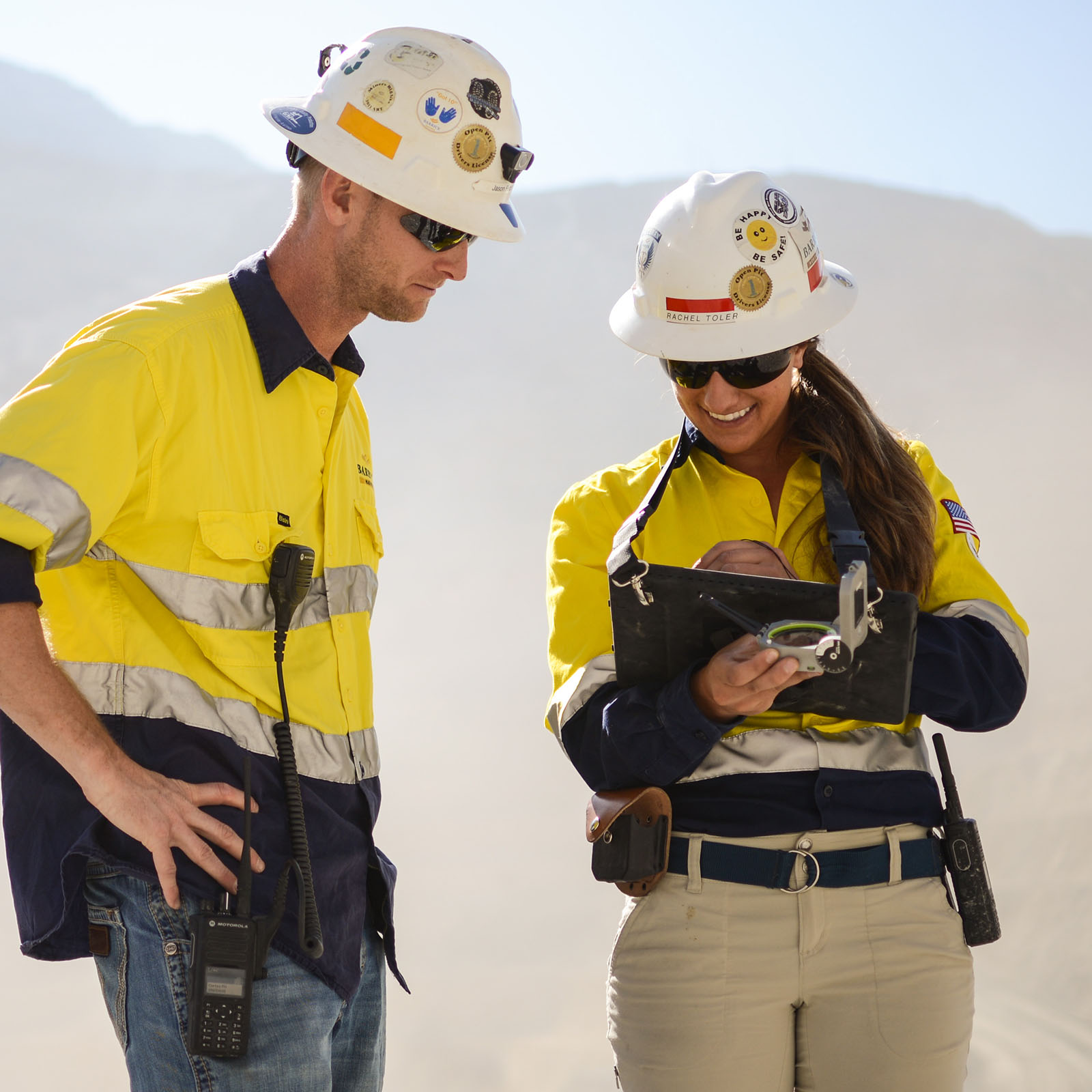 Two mining workers in high-visibility yellow safety shirts and white hard hats decorated with various stickers stand outdoors against a desert mountain backdrop. One worker carries a radio on their belt while the other smiles while looking at a tablet. Both are wearing safety gear including sunglasses, and their hard hats display company stickers and safety badges.