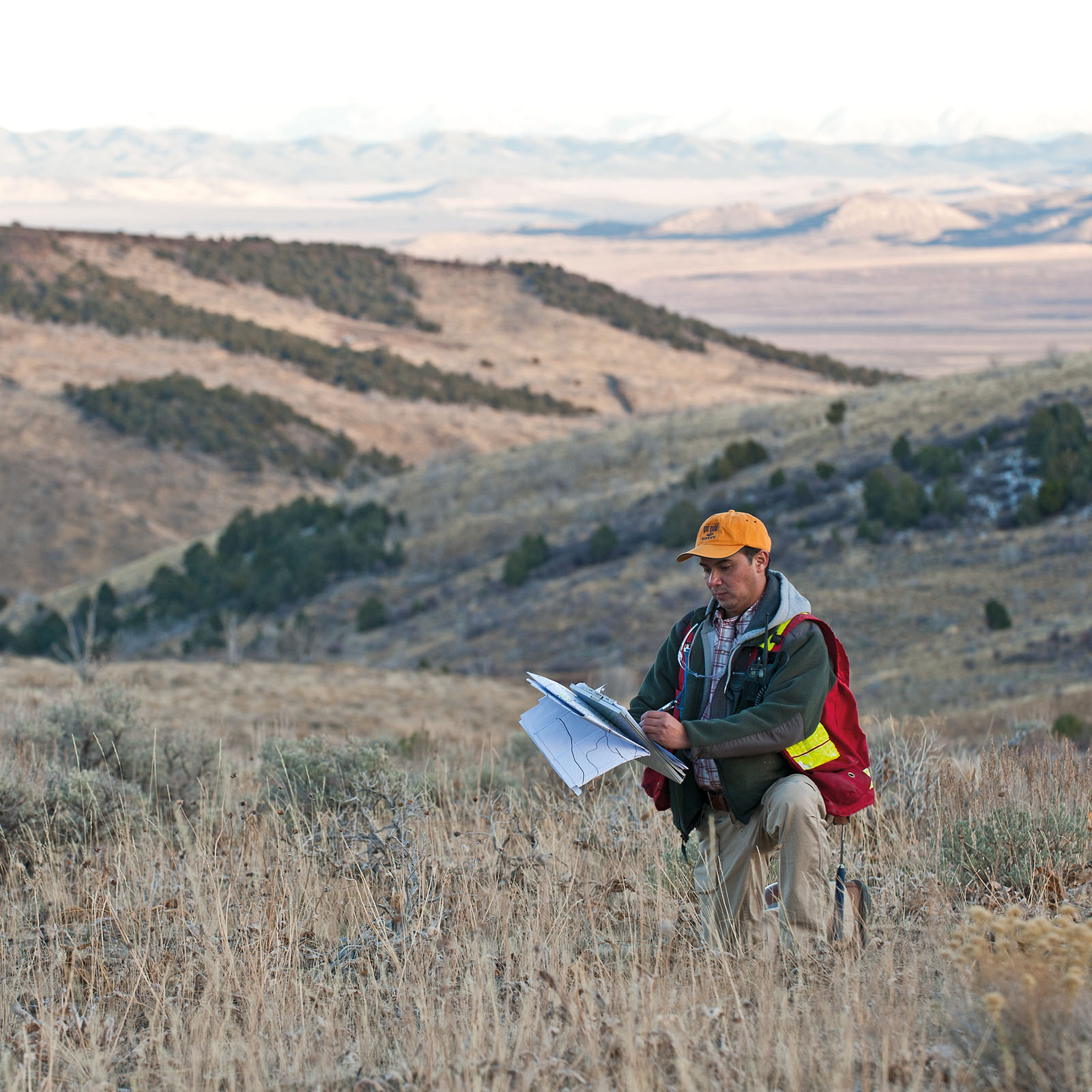 A person in field work attire — wearing an orange baseball cap, green jacket with reflective strips, and khaki pants — examines documents on a clipboard while standing in dry grassland. They carry a red backpack and are positioned on a hillside with rolling mountains and desert landscape visible in the background.