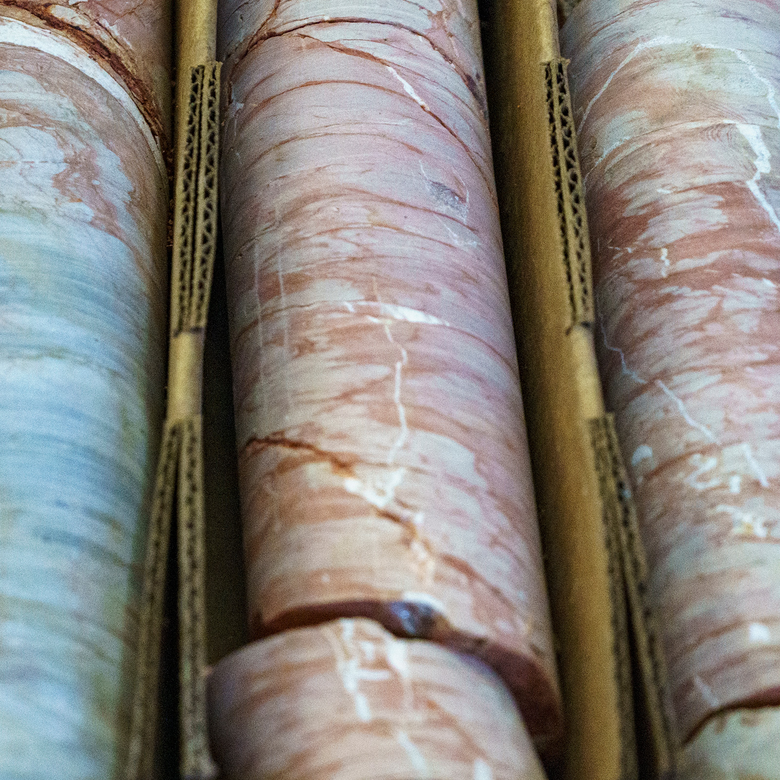 Close-up photograph of drill core samples arranged in parallel rows. The cores show pink and white marbled rock patterns with visible striations and veining. They are stored in standard core boxes with dividers between each core section.