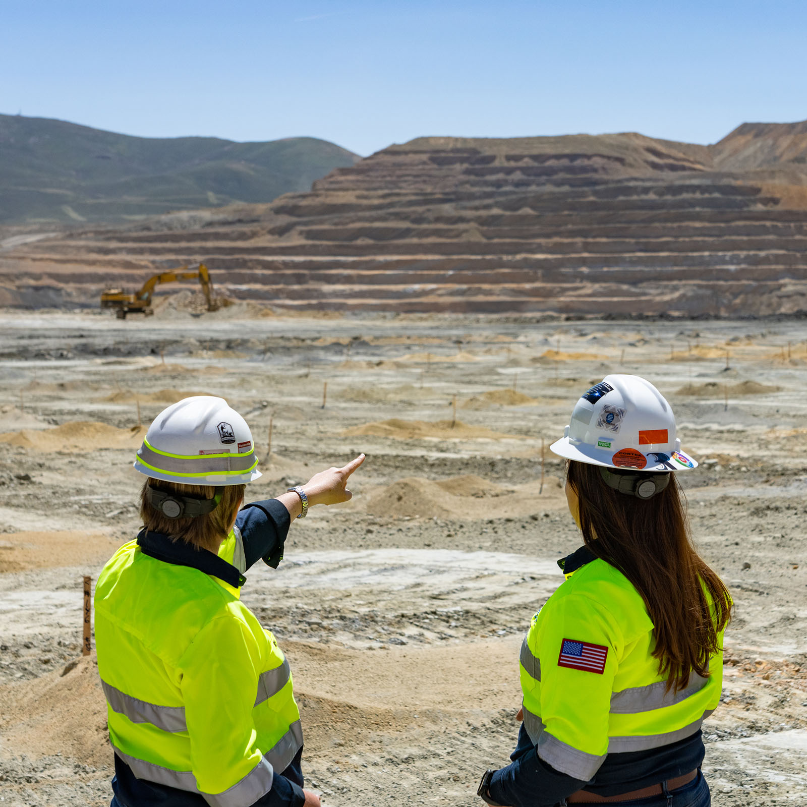 Two mining engineers wearing white hard hats and high-visibility yellow safety vests stand on a ledge overlooking an open pit mine. One engineer points toward a yellow excavator working on the terraced mining site in the distance. The backdrop shows stepped layers of exposed earth and hills.