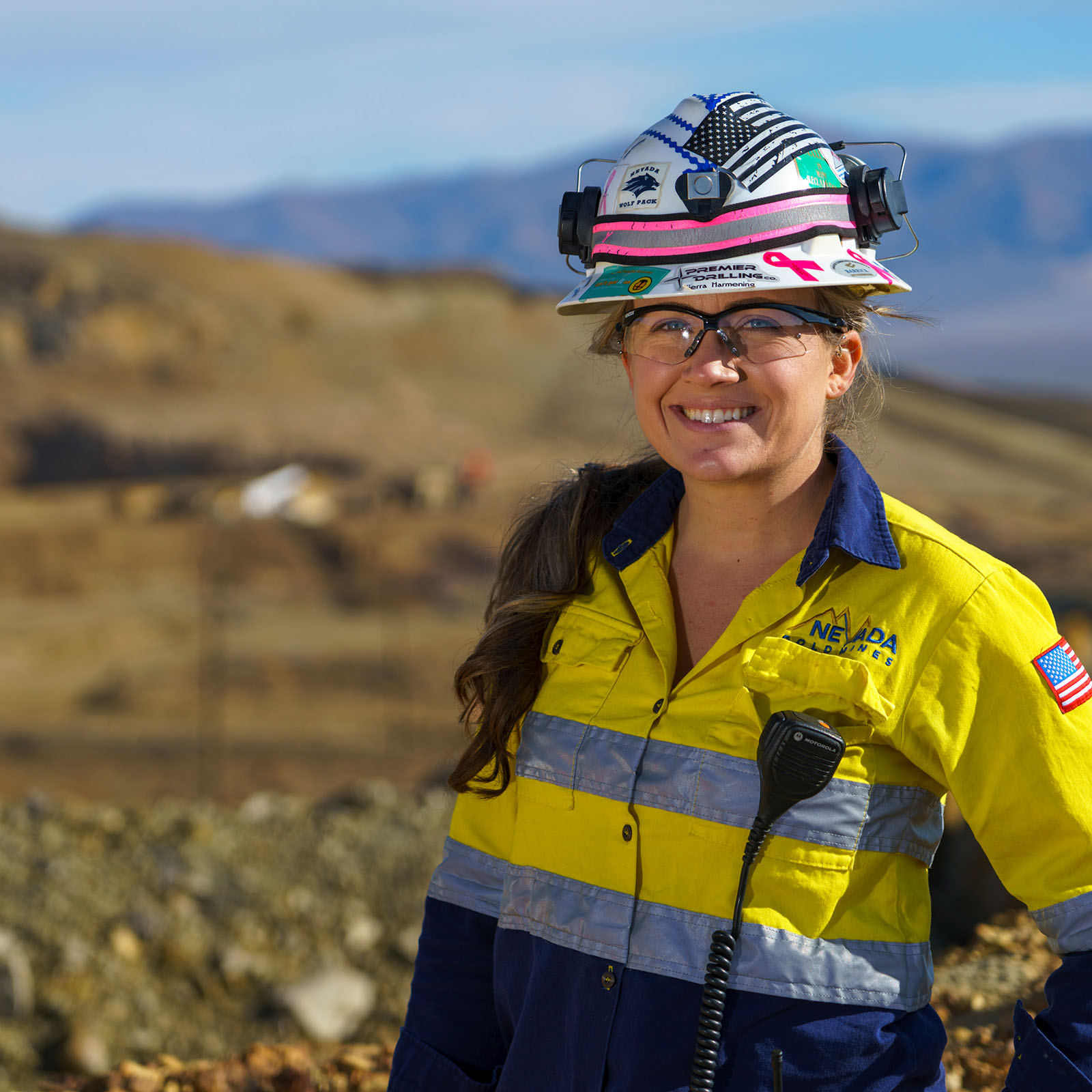 A smiling female mining employee wearing safety gear including a decorated hard hat with American flag design and pink stripe, safety glasses, and a high-visibility yellow and navy Nevada Gold Mines uniform with reflective stripes. She has a radio attached to her uniform and stands outdoors with desert hills in the background.
