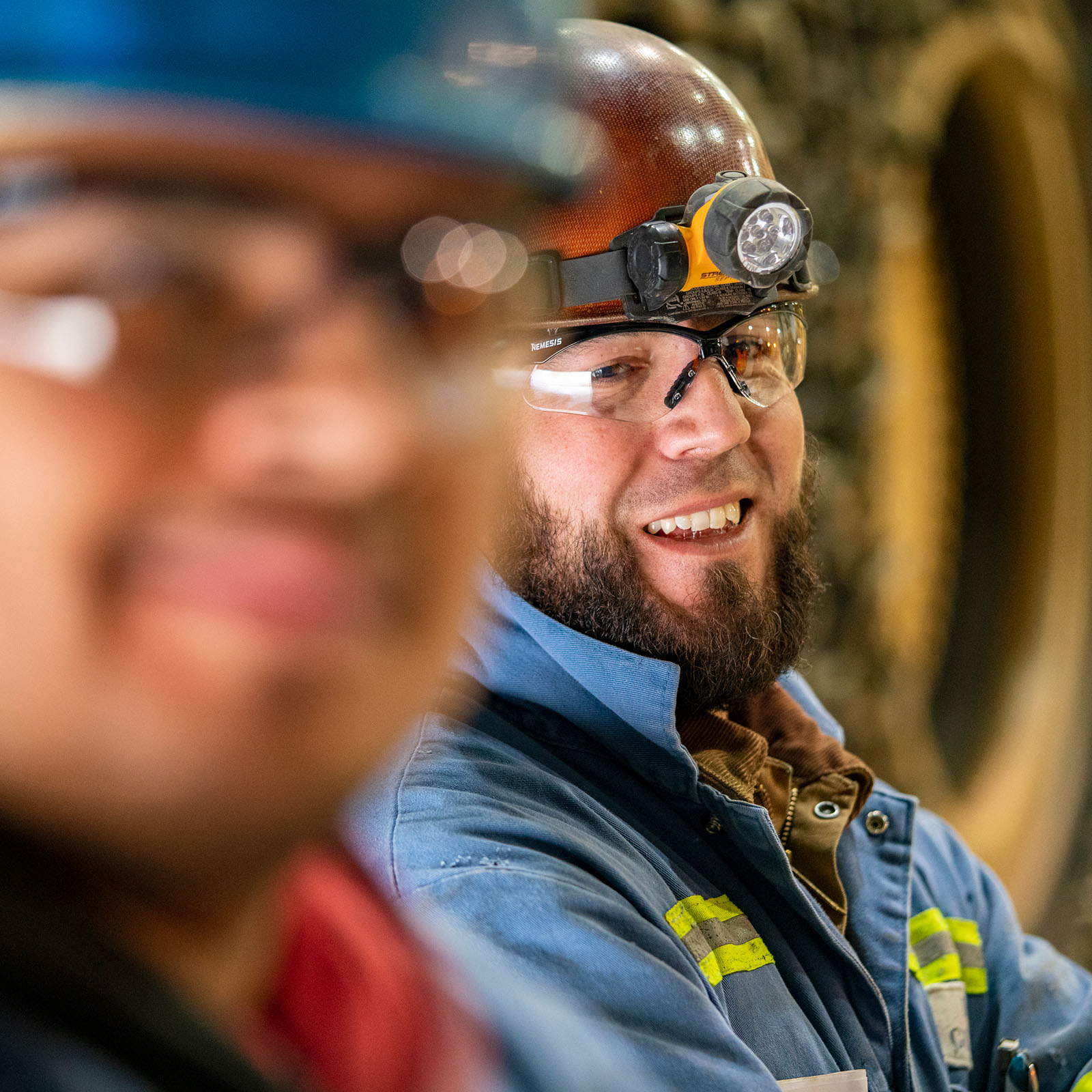 A smiling mine worker wearing safety equipment including a hard hat with a mounted LED headlamp, protective safety glasses, and a blue work uniform with reflective stripes. The person has a full dark beard and is shown in profile, with another worker blurred in the foreground.