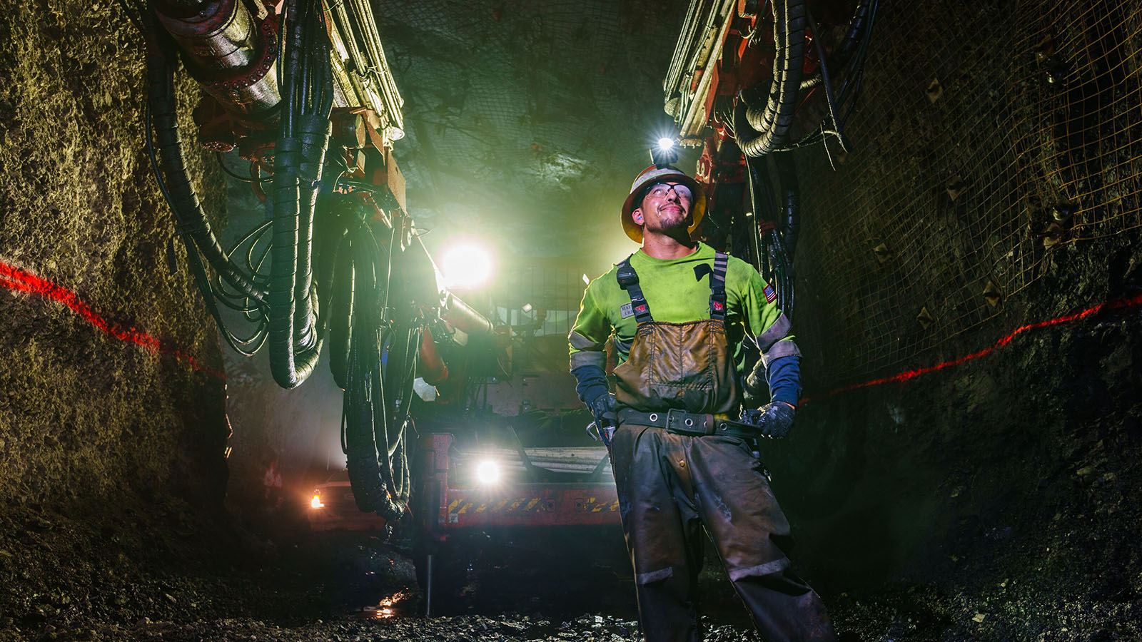 A miner in high-visibility gear stands in a dimly lit underground tunnel at the Goldrush mine in Nevada, surrounded by heavy drilling machinery and illuminated by bright work lights against the rocky walls and wet ground.